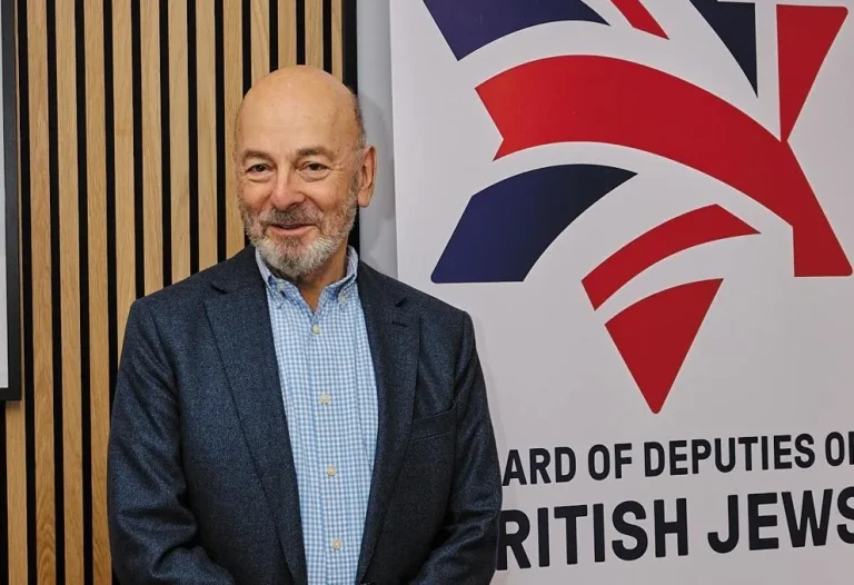 A Photo of Vivian Wineman standing in front of a Board of Deputies banner. Vivian is wearing a navy blue shit jacket and a blue and white check shirt.